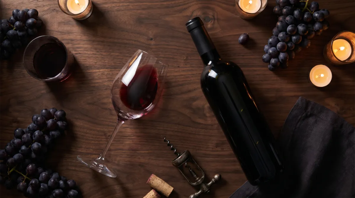A bottle of red wine and a glass on a dark table with grapes and a corkscrew